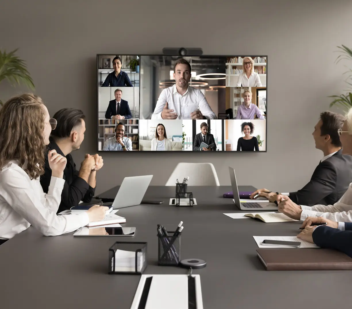A group of people sitting at a table and viewing a virtual meeting on a screen mounted to the wall.